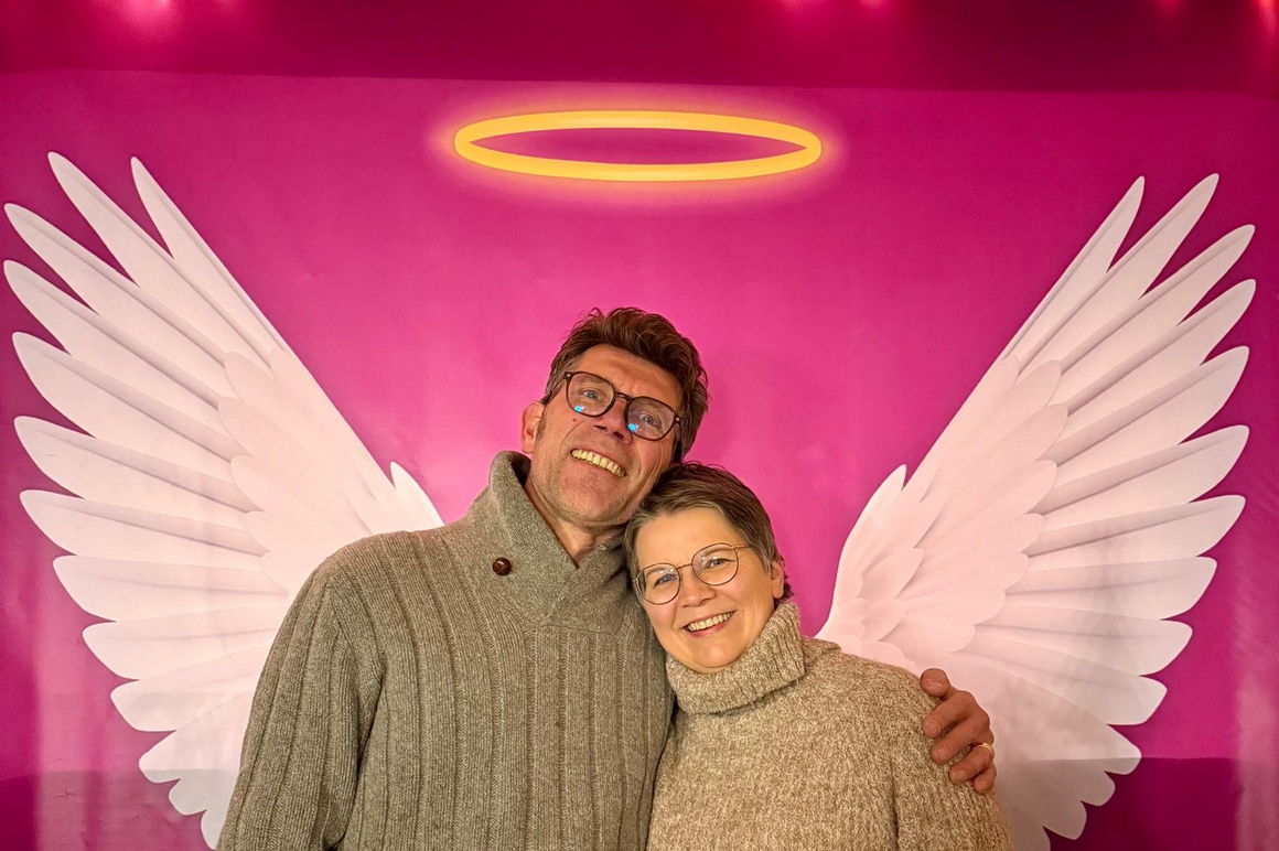 Eine männlich und eine weiblich gelesene Person halten sich im Arm, hinter ihnen eine Wand, auf der Engelsflügel und ein Heiligenschein gemalt sind.