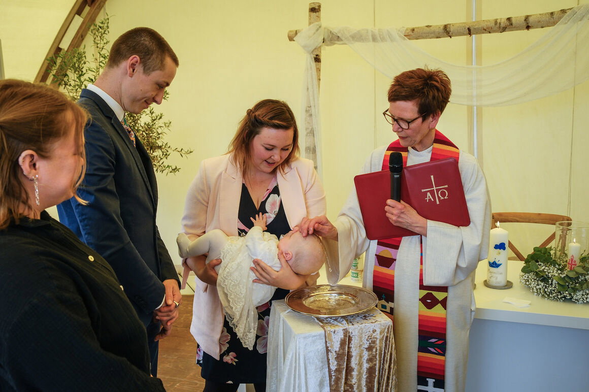 Eine Pastorin tauft in einem Zelt ein Baby, das die Mutter über die Taufschale hält.