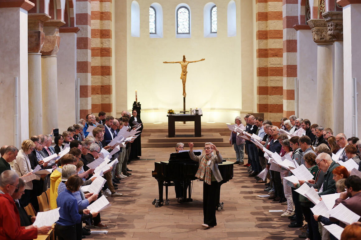 Viele Menschen singen in einer Kirche. Eine als Frau lesbare Person dirigiert, eine andere spielt am Flügel.